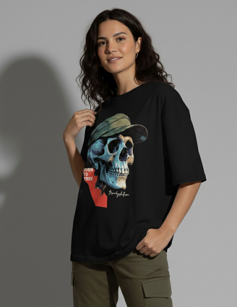 A young woman with dark, wavy hair stands facing slightly to the right, looking at the camera with a gentle smile. She wears a black crew-neck t-shirt with a large graphic of a blue and beige skull wearing a green cap and the text "BORN TO TREK". Her left hand is casually in her olive green cargo pant pocket, while her right hand lightly touches the collar of her t-shirt. A shadow is cast behind her on a light gray background. The Smoky Skull