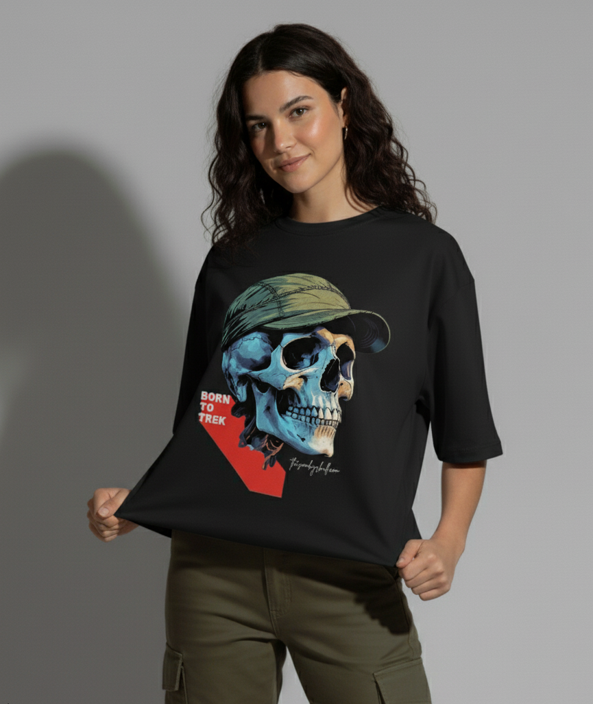 A young woman with dark, wavy hair stands facing slightly to the right, smiling at the camera. She is wearing a black crew-neck t-shirt with a large graphic of a blue and beige skull wearing a green cap and the text "BORN TO TREK". She holds the bottom of the t-shirt with both hands, pulling it slightly to display the design. She wears olive green cargo pants, and a shadow is cast behind her on a light gray background. The Smoky Skull