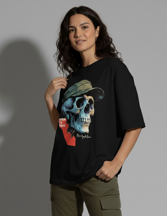 A young woman with dark, wavy hair stands facing slightly to the right, looking at the camera with a gentle smile. She wears a black crew-neck t-shirt with a large graphic of a blue and beige skull wearing a green cap and the text "BORN TO TREK". Her left hand is casually in her olive green cargo pant pocket, while her right hand lightly touches the collar of her t-shirt. A shadow is cast behind her on a light gray background. The Smoky Skull