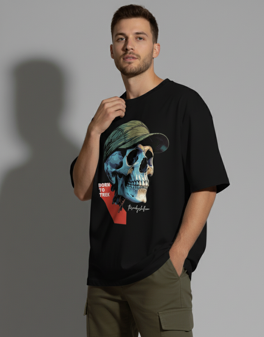 A young man with short dark hair and a light beard stands facing slightly to the right, looking at the camera with a confident expression. He wears a black crew-neck t-shirt with a large graphic of a blue and beige skull wearing a green cap and the text "BORN TO TREK". His right hand is casually in his olive green cargo pant pocket, while his left hand lightly touches the collar of his t-shirt. A shadow is cast behind him on a light gray background. The Smoky Skull