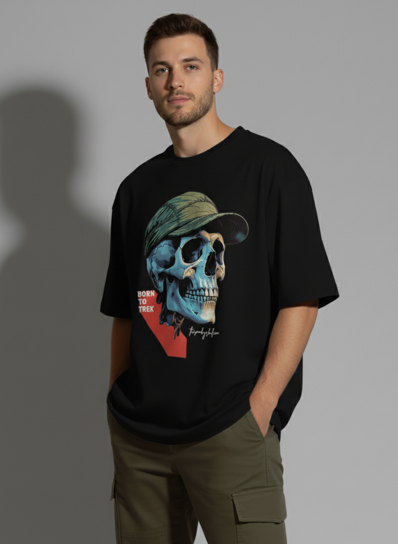 A young man with short dark hair and a light beard stands facing slightly to the right, looking at the camera with a serious expression. He wears a black crew-neck t-shirt with a large graphic of a blue and beige skull wearing a green cap and the text "BORN TO TREK". Both of his hands are casually in the pockets of his olive green cargo pants. A shadow is cast behind him on a light gray background. The Smoky Skull