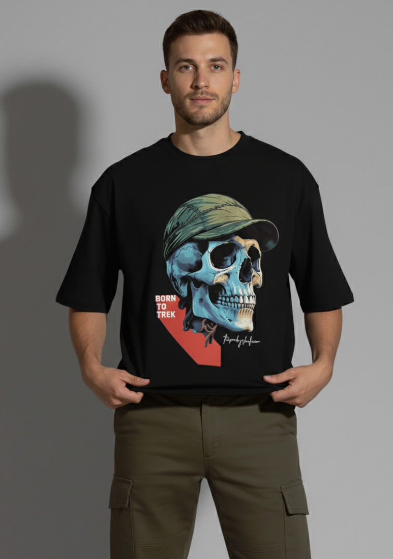  A young man with short dark hair and a light beard stands facing forward, looking at the camera with a direct gaze. He is wearing a black crew-neck t-shirt with a large graphic of a blue and beige skull wearing a green cap and the text "BORN TO TREK". He holds the bottom of the t-shirt with both hands, pulling it slightly to display the design. He wears olive green cargo pants, and a shadow is cast behind him on a light gray background. The Smoky Skull